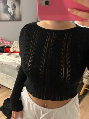 Svart croppad stickad tröja - Säljer en svart croppad stickad tröja med lång ärm och snyggt hålmönster. Tröjan har rund halsringning och sitter tajt men bekvämt. Perfekt att styla med höga jeans eller kjol för en trendig look.