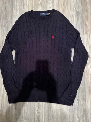 Mörkblå kabelstickad tröja Polo Ralph Lauren - Kabelstickad mörkblå tröja från Polo Ralph Lauren med klassisk röd broderad logga på bröstet. Tröjan har rund halsringning och långa ärmar, perfekt för lager på lager under höst och vinter.