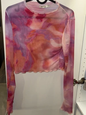Mesh topp i rosa och lila från Nelly - Säljer en festlig mesh topp från Nelly i storlek S. Toppen har ett abstrakt mönster i rosa, lila och orange toner, är långärmad och croppad med vågig kant nertill. 