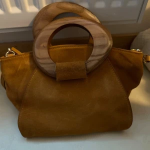 Mocka handväska från Mango - Säljer en snygg handväska från Mango i mocka med unika runda handtag i trä. Väskan har avtagbar axelrem och dragkedja upptill. Insidan är svart med en innerficka. Märkena som kan ses på väskan från handtagen kan enkelt borstas bort på grund av mocka materialet.