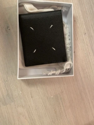 Maison Margiela svart plånbok i skinn - Maison Margiela plånbok i svart skinn med de ikoniska fyra vita sömmarna på framsidan. Klassisk och stilren design med flera fack för kort och sedlar. Kommer med original dustbag och box. Perfekt för dig som gillar minimalistisk och lyxig stil.