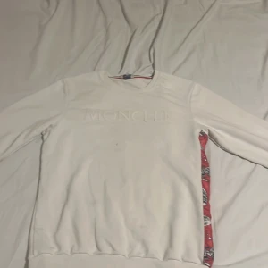 Vit Moncler sweatshirt med röd detalj - Vit sweatshirt från Moncler med präglad logga framtill och unik präglad design på ryggen. Sidorna har röda paneler med tecknade tryck. Tröjan har rund halsringning och långa ärmar med ribbade muddar. Perfekt för dig som gillar streetwear med exklusiv känsla.
