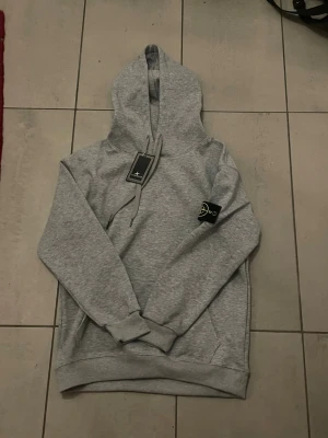 Stone Island hoodie - Säljer en grå stone island hoodie i nytt skick. Storleken är L i barn storlek som ungefär motsvarar xs i vuxen.