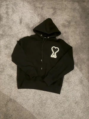 Svart hoodie från Ami Paris - Säljer en svart hoodie från Ami Paris med stor vit hjärtdetalj och bokstaven A på bröstet. Tröjan har huva med snörning, känguruficka och ribbade muddar. Tillverkad i mjuk bomull, perfekt för en chill och stilren look.