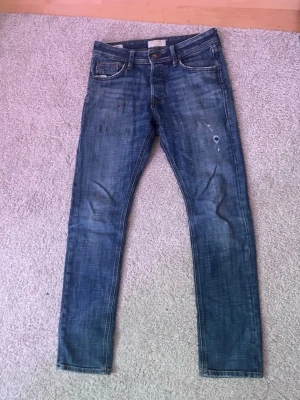 Blå skinny jeans från Jack & Jones - Säljer ett par blå skinny jeans från Jack & Jones med klassisk femficksdesign och slitna detaljer på benen. Jeansen har normal midja och är tillverkade i mjukt denimtyg med snygg tvätt och kontrastsömmar. Perfekta för en avslappnad och trendig look.