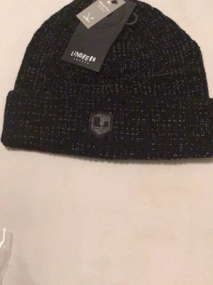 Svart stickad mössa från Lindberg - Säljer en svart stickad mössa från Lindberg med diskret logga framtill och reflekterande detaljer. Mössan har en klassisk ribbad kant och är tillverkad i mjukt material som passar perfekt för kyliga dagar.