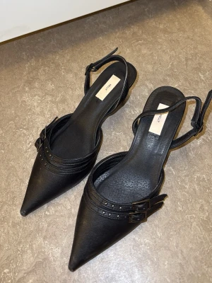 Svarta pumps med spetsig tå från Nelly - Säljer ett par trendiga svarta pumps från Nelly med spetsig tå och dubbla spännen på sidan. 