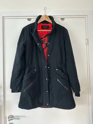 Svart  jacka från Sisters Point - Svart jacka från Sisters Point med röd innerfoder. Jackan har långa ärmar, dragkedja och knappar framtill samt två snedställda fickor med dragkedja. Midjan är lätt markerad med resår och jackan har en klassisk hög krage.