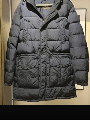 Mörkblå dunjacka från Oscar Jacobson - Snygg mörkblå dunjacka från Oscar Jacobson med huva, stora fickor med knappar och både dragkedja och knappstängning. Jackan har quiltad design och lång passform som håller dig varm under kalla dagar. Perfekt för dig som vill ha både stil och funktion.