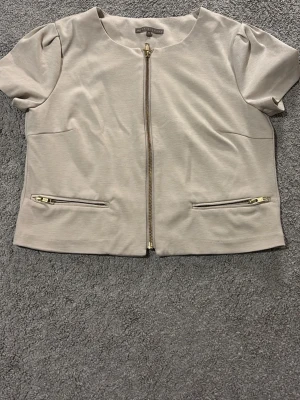 Beige overshirt från mint&berry - Kortärmad beige overshirt från mint&berry i mjukt material. Jackan har rund hals, raka linjer och dragkedja framtill samt två snedställda fickor med guldfärgade dragkedjor. Perfekt för en clean och stilren look.