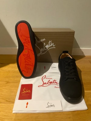 Christian Louboutin skor Helt Nya!!! - Säljer ett par svarta sneakers från Christian Louboutin med ikonisk röd sula. Skorna har rund tå, snörning och är tillverkade i skinn med mattsvart finish. Kommer med originalkartong, dustbag och äkthetskort. Perfekta för dig som vill ha något extra till din outfit.                                                                         Helt Nya!!!