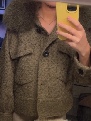 Beige jacka med päls från Meotine XS/S - Säljer en khaki färgad jacka från Meotine i storlek XS/S. Jackan har en stor fluffig pälskrage, två stora fickor framtill och stängs med knappar. Materialet är ullblandning med struktur och den har en boxig, lite kortare passform. Perfekt för dig som vill sticka ut i höst och vinter! Kan gå ner i pris vid snabb affär!!