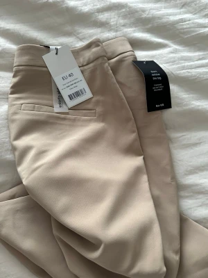 Beige kostymbyxor från NA-KD med slits - Snygga beige kostymbyxor från NA-KD i rak modell med hög midja och slits framtill vid benslutet. Byxorna har en clean look med diskreta fickor bak och är gjorda i ett mjukt material som känns lyxigt. Perfekta att styla med både boots och klackar.