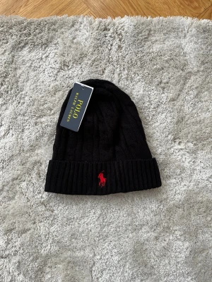 Svart stickad mössa från Polo Ralph Lauren - Säljer en svart stickad mössa från Polo Ralph Lauren med klassisk kabelstickning och uppvikt kant. Mössan har en röd broderad logga framtill och är tillverkad i mjukt material som håller dig varm under kalla dagar.