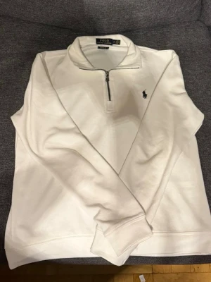 Vit half zip tröja från Polo Ralph Lauren - Säljer en stilren vit tröja från Polo Ralph Lauren i bomull med half zip och klassisk krage. Tröjan har lång ärm och den ikoniska loggan broderad på bröstet. Perfekt för dig som gillar enkel och clean stil.