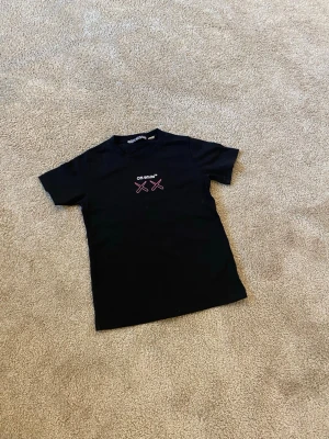 Svart Off-White t-shirt med tryck - Säljer en svart t-shirt från Off-White med ikoniskt tryck framtill och stort grafiskt motiv på ryggen i vitt, rosa och lila. Klassisk rund hals och korta ärmar. Materialet är mjuk bomull som känns skön mot huden. Perfekt för dig som gillar streetwear och vill sticka ut.