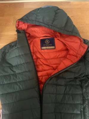 Grön pufferjacka från Henri Lloyd - Säljer en grön pufferjacka från Henri Lloyd med orange foder och huva. Jackan har quiltad design, dragkedja framtill och en broderad logga på bröstet. Perfekt för kyliga dagar och riktigt snygg färgkombo!