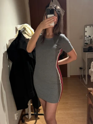 Grå tight kortklänning med ränder - Säljer en grå tight kortklänning från H&M med vita och röda ränder längs sidorna. Klänningen har rund halsringning och korta ärmar. Super bra skick!