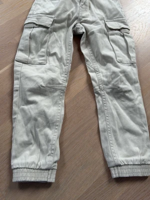 Beiga cargopants från 157, storlek 100 - Säljer ett par beiga cargopants från 157 i storlek 100. Byxorna har stora sidofickor, resår i midjan och vid bensluten samt justerbar midja med knapp. Tillverkade i mjuk bomull för skön passform.
