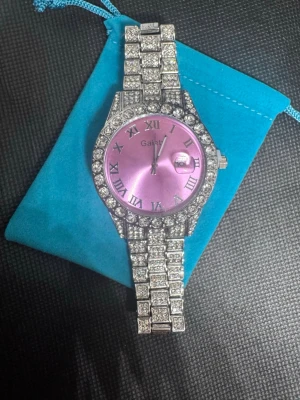 Lila glittrig klocka från Gaiety - Säljer en riktigt blingig klocka från Gaiety med lila urtavla och romerska siffror. Armbandet och boetten är täckta av gnistrande stenar som ger maxad glamour. Klockan har datumvisning och ett rejält metallarmband. Perfekt för dig som vill sticka ut!