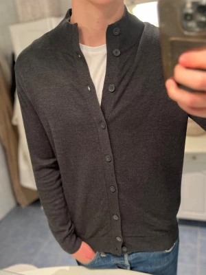 Mörkgrå kofta med knappar - Snygg mörkgrå kofta med raka linjer och klassisk knappstängning hela vägen. Tröjan har lång ärm och ribbad krage som ger en clean look. Perfekt att slänga över en t-shirt för en chill men ändå stilren outfit.