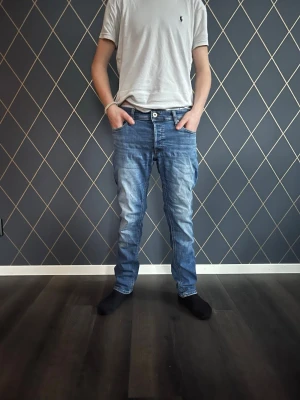 Blå jeans från Jack & Jones - Säljer ett par klassiska blå jeans från Jack & Jones med normal passform och raka ben. Jeansen har fem fickor, bälteshällor och snygga slitningar framtill för en avslappnad look. Tillverkade i mjukt denimtyg som är skönt att bära hela dagen. Pris kan diskuteras vid snabb affär! Modellen är 165cm. Taggen säger 30/31 men är lite mindre så passar runt 160/170