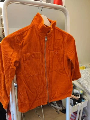 Orange manchesterjacka från Arket - Säljer en riktigt snygg orange manchesterjacka från Arket i ekologisk bomull. Jackan har två stora bröstfickor med knappar, hög krage och dragkedja framtill. Perfekt för dig som vill sticka ut med färg och cool retrostil.