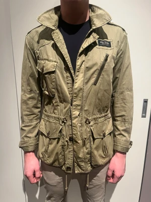 Olivgrön parkajacka med snörning - Säljer en olivgrön parkajacka från Morris med flera praktiska fickor, snörning i midjan och dragkedja framtill. Jackan har hög krage och klassisk militärinspirerad look. Perfekt för dig som gillar streetstyle och funktionella detaljer.