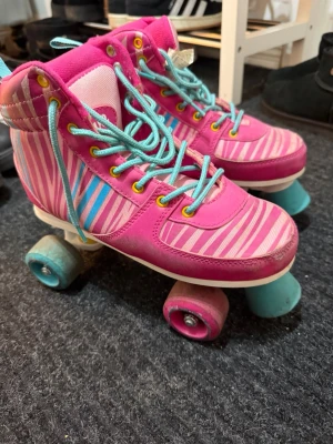  rullskridskor - Säljer ett par färgglada rullskridskor från Rookie med rosa och ljusblå ränder, turkosa skosnören och gula öljetter. Skorna har högt skaft och hjul i rosa och blått. Perfekta för dig som vill sticka ut på rinken!