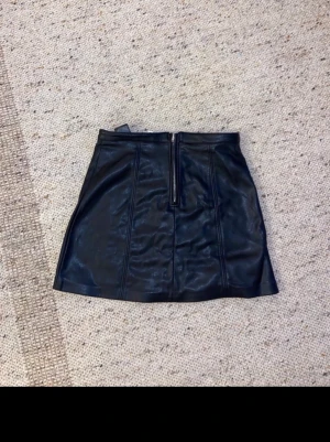 Svart kortkjol i skinnimitation Divided helt ny  - Säljer en svart kortkjol från Divided i skinnimitation med markerade sömmar och dragkedja framtill. Kjolen har en clean och edgy look, perfekt för dig som gillar att sticka ut. Materialet är polyester med polyuretanbeläggning.