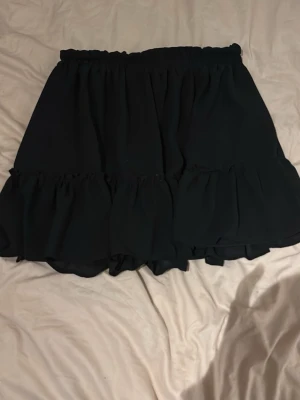 Svart volangkjol med resårmidja - Supersnygg svart kortkjol med volangdetaljer och resår i midjan. Kjolen har en luftig och flowy känsla, perfekt för dig som gillar en enkel men ändå trendig stil. Passar till många olika looks och är lätt att matcha.