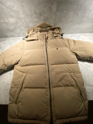 Beige pufferjacka Polo Ralph Lauren S - Säljer en beige pufferjacka från Polo Ralph Lauren i storlek S. Jackan har huva, två fickor med dragkedja och klassisk Polo-logga på bröstet. Perfekt för kalla dagar och riktigt skön att bära. Stängs med grov dragkedja framtill. Är 156 men passar 160