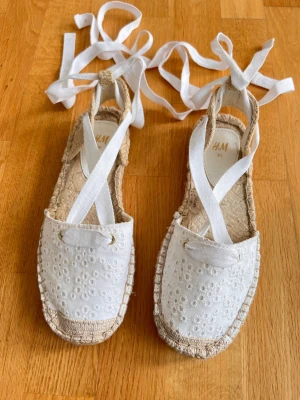 Snygga sandaler från H&M - H&M. Storlek 36.    i storlek 36 med vit broderad ovandel i bomull och långa band att knyta runt ankeln. Sula i naturfärgat flätat material ger en boho-vibe. Perfekta för varma dagar och sommaroutfits!
