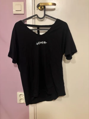 Svart t-shirt med öppen rygg från Remina - Svart t-shirt från Remina i storlek S. Framsidan har trycket 'LOVER' med ett streck över. T-shirten har korta ärmar och en djup, öppen rygg med dekorativa band. Tillverkad i mjukt bomullstyg och har en loose passform.