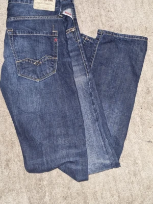 Replay blå jeans, rak passform - Säljer ett par klassiska blå jeans från Replay med raka ben och snygga slitningar. Jeansen har kontrastsömmar, fem fickor och en röd detalj vid hällan. Märkespatch baktill och tydlig Replay-logga. Perfekta till sneakers eller boots.