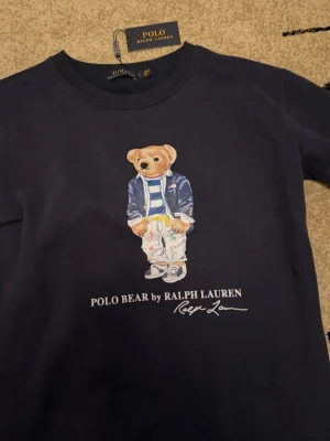 Mörkblå långärmad Polo Bear-tröja - Snygg mörkblå långärmad tröja från Polo Ralph Lauren med ikoniska Polo Bear-trycket på bröstet. Rund halsringning och klassisk passform. Tillverkad i mjuk bomull som är skön att ha på sig. Perfekt för dig som gillar streetstyle och vill sticka ut lite extra.