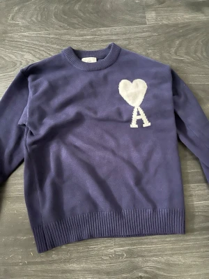 Mörkblå stickad tröja från Ami Paris  - Säljer en mörkblå stickad tröja från Ami Alexandre Mattiussi med vit hjärt- och A-detalj på bröstet. Tröjan har rund hals, ribbade muddar och är långärmad. Perfekt för dig som gillar stilrena plagg med en twist. PRIS ÄR INTE FAST!