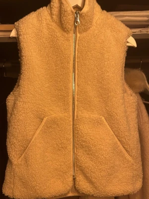 Beige teddyväst med dragkedja - Mysig beige teddyväst med hög krage och två stora fickor framtill. Västen har en rak passform och stängs med en silvrig dragkedja. Perfekt att slänga över en hoodie eller tröja för extra värme och stil.