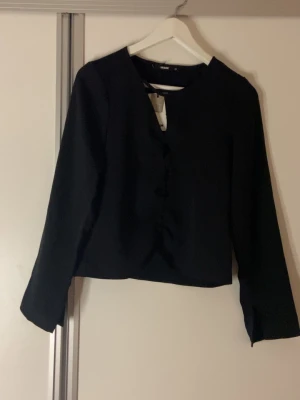 Svart omlottblus från Bik Bok - Snygg svart omlottblus från Bik Bok i blankt polyestertyg. Blusen har långa ärmar och en twistad osymetrisk detalj framtill som ger en cool look. 