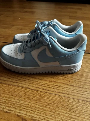 Nike Air Force 1 ljusblå och vita - Säljer ett par Nike Air Force 1 i en fräsch ljusblå och vit färgkombination. Skorna har klassisk låg siluett, snörning och perforerad tå för extra ventilation. Tillverkade i en mix av skinn och syntetmaterial med vit sula och broderad Nike-logga på hälen. Använd fåtal gånger 