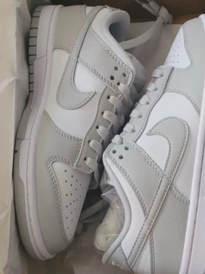Nike Dunk Low vit/grå sneakers - Säljer ett par Nike Dunk Low sneakers i vitt och ljusgrått. Skorna har klassisk låg siluett, snörning och den ikoniska swoosh-loggan på sidan. Ovandelen är i slätt skinn och syntet, med perforerade detaljer på tån för extra ventilation. Perfekta för dig som gillar stilrena och tidlösa sneakers.