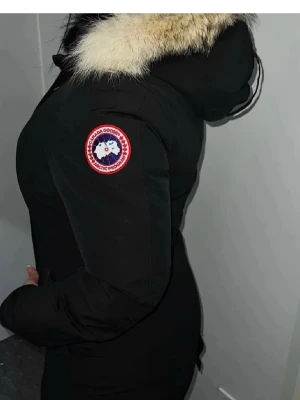 Svart parkas från Canada Goose - Säljer en svart parkas från Canada Goose med pälskantad huva och klassisk logga på ärmen. Jackan har långa ärmar, knappar framtill och stora fickor. Perfekt för kalla dagar och riktigt snygg passform.XSS/ XS