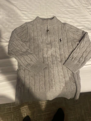 Grå kabelstickad tröja Polo Ralph Lauren - Säljer en grå kabelstickad tröja från Polo Ralph Lauren i storlek S. Tröjan har half zip, ribbade muddar och hög krage. Klassisk logga broderad på bröstet och riktigt skön passform. Perfekt till kyliga dagar!