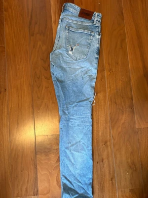 Jeans Tommy Hilfiger - Säljer ett par ljusblå skinny jeans från Tommy Hilfiger med slitningar och hål på ena benet. Klassisk femficksmodell med läderpatch bak i midjan och diskret logga på bakfickan. Jeansen har normal passform och är tillverkade i bomull.