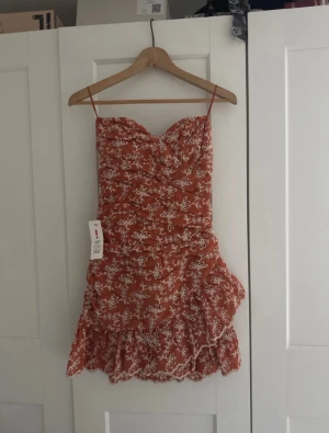 Kort klänning med volang - Supersöt kort klänning i rostbrunt med vita broderade blommor. Volangkant nertill och smickrande rynkningar. Dragkedja i ryggen. Perfekt till sommaren!❤️ Strl XS. Aldrig använd med alla lappar kvar. Ca 1500kr nypris.