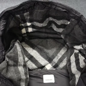 Svart dunjacka från Burberry - Säljer en svart dunjacka från Burberry med klassisk logotyp på ärmen och rutigt foder i grått och vitt. Jackan har huva, dragkedja och knappar framtill samt praktiska fickor. Perfekt för kalla dagar med sin quiltade design och varma material.