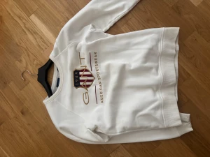 Vit sweatshirt från GANT - Säljer en fräsch vit sweatshirt från GANT med tryckt logga och emblem i guld, blått och rött på bröstet. Tröjan har rund hals, ribbade muddar och är långärmad. Perfekt för en sportig och clean look.