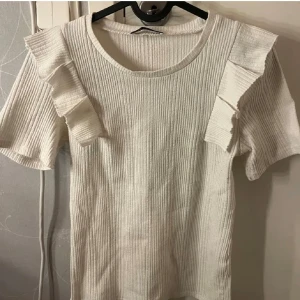 Vit ribbad topp med volang från Zara - Säljer en vit ribbad topp från Zara i storlek S. Toppen har korta ärmar och snygga volangdetaljer på axlarna. Perfekt för dig som gillar stilrena och trendiga plagg.