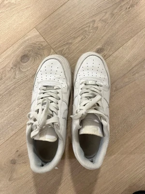 Vita Nike Air Force 1 sneakers - Säljer ett par klassiska vita Nike Air Force 1 sneakers med snörning och perforerad tå. Skorna har en platt sula och rund tå, tillverkade i skinn med textilfoder. Perfekt för dig som gillar tidlös streetstil och vill ha ett par ikoniska sneakers i garderoben.