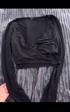 Svart omlottopp med v-ringning - Säljer en svart omlottopp med lång ärm och djup v-ringning. Toppen är croppad och har en stilren look som passar perfekt till höga jeans eller kjol. Materialet känns mjukt och stretchigt, vilket gör den bekväm att bära. Perfekt för dig som gillar minimalistisk stil.